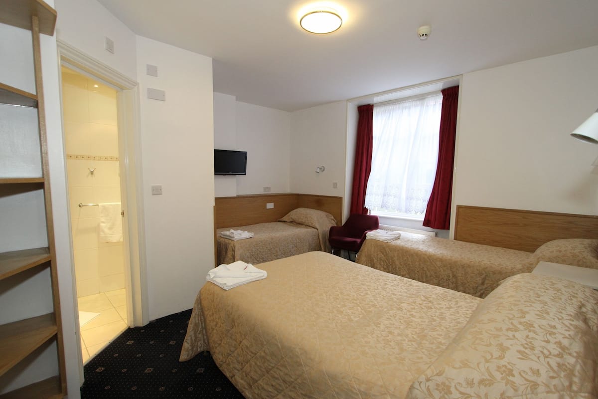 Habitación de hotel con dos camas individuales, cabecero de madera, televisor en la pared, ventana con cortinas rojas y puerta que da a un baño.