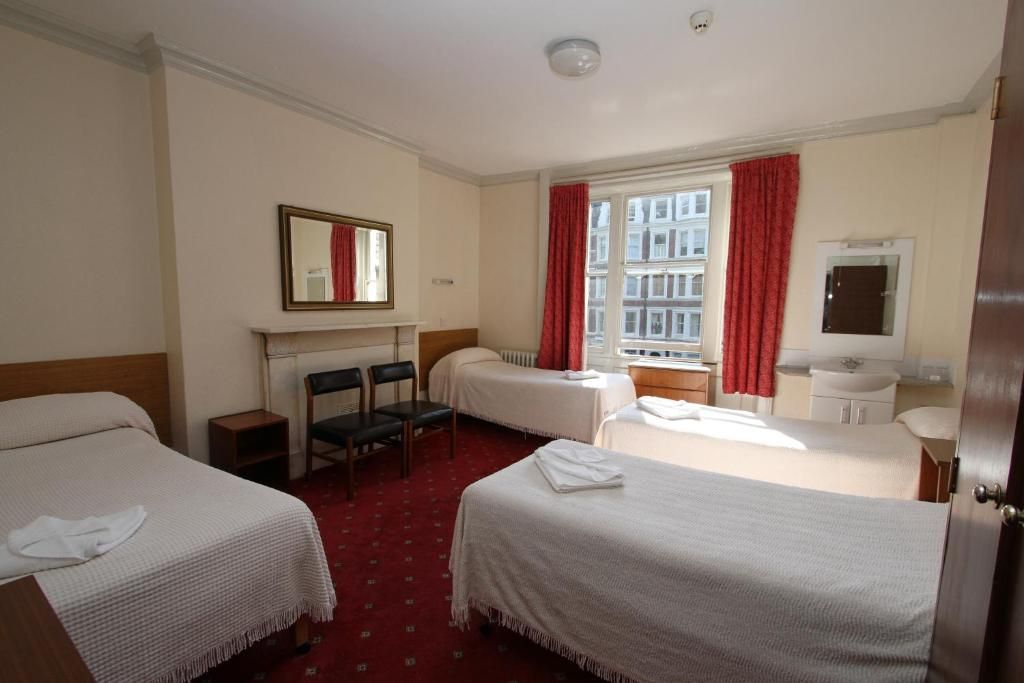 A hotel room with two single beds, a small table, a chair, a mirror, a window with red curtains, and a radiator.