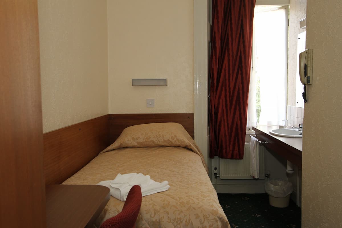 Una habitación pequeña con una cama individual, cabecera de madera, mesita de noche, ventana con cortinas rojas, baño con lavamanos y espejo, y un radiador.