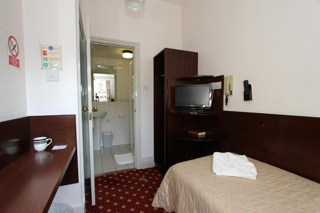 Une chambre d’hôtel avec un lit, un téléviseur posé sur un meuble, un bureau avec une tasse, et une porte donnant accès à une salle de bain.