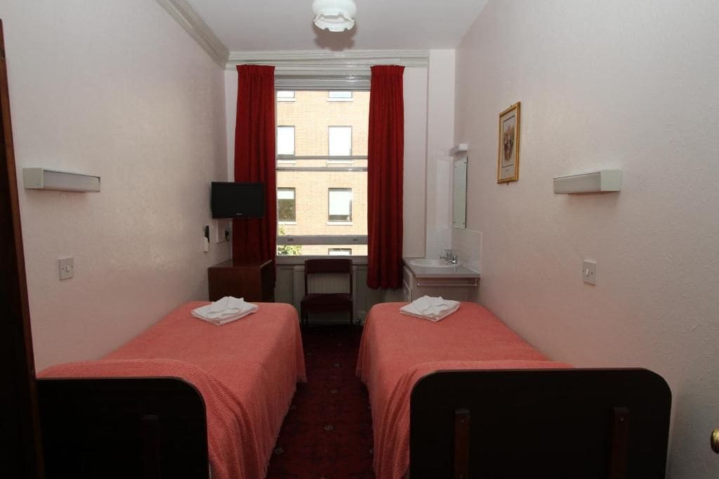 Habitación de hotel con dos camas individuales, un escritorio con silla, televisor y ventana con cortinas rojas, con vistas a edificios.