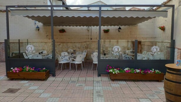 An outdoor seating area with tables and chairs, enclosed by a glass partition, and decorated with flower planters.
