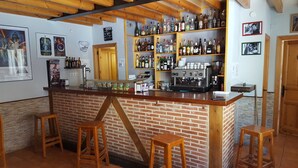 A bar area with a brick counter, stools, shelves with bottles, a coffee machine, and framed pictures on the wall.