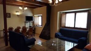 A living room with a blue sofa, a glass coffee table, wooden chairs, a television, a fireplace, and a window with curtains.