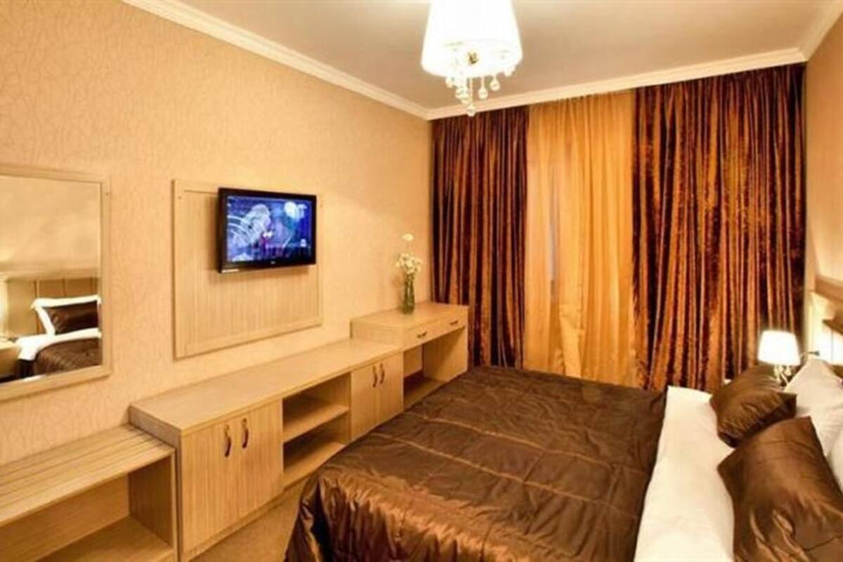 A hotel room with a bed, a TV mounted on the wall, a vanity with a mirror, and a large window with curtains.