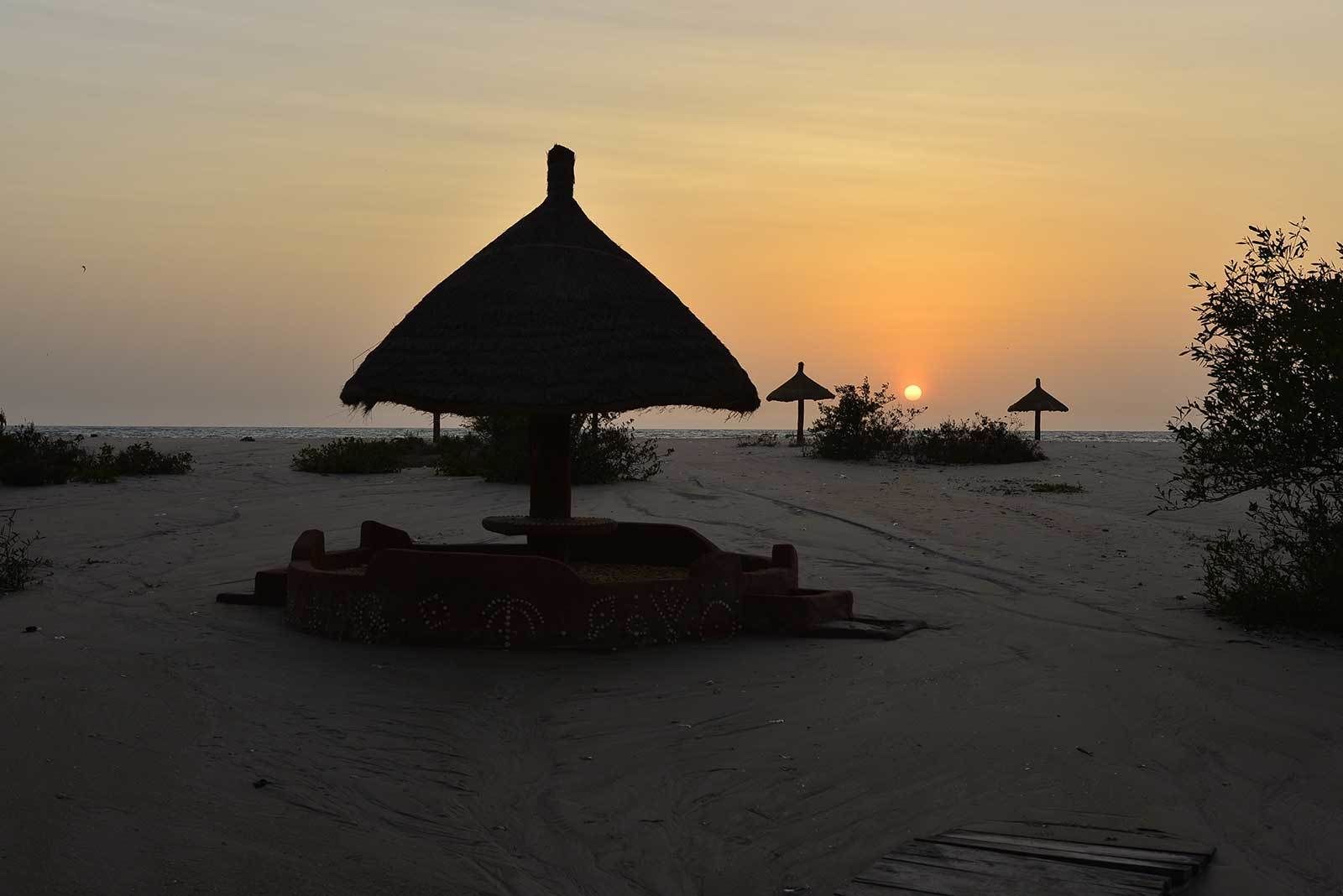 A beach scene with a thatched umbrella structure, a small table, and a bench.
