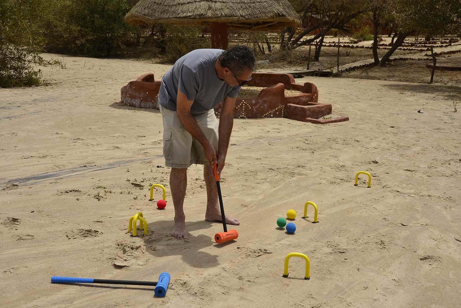 A person playing croquet on a sandy field with a thatched-roof pavilion in the background.
