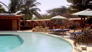 A swimming pool with lounge chairs and umbrellas, surrounded by palm trees and a building with arched windows. - Ecolodge de Palmarin (Fatick)