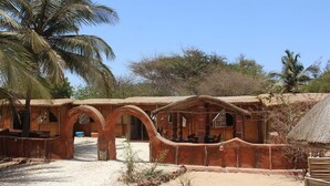 A rustic building with arched doorways and a thatched roof, surrounded by palm trees and dry vegetation. - Ecolodge de Palmarin (Fatick)