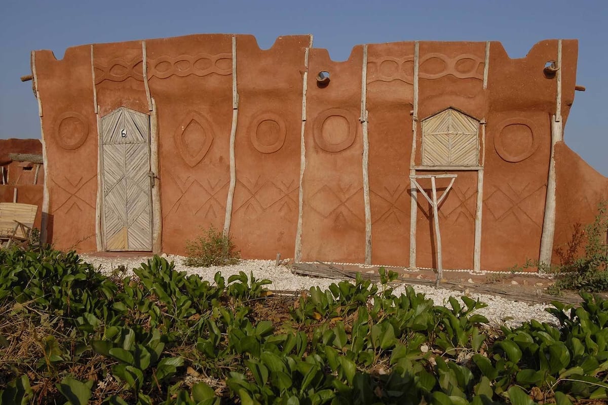 A traditional mud-brick building with geometric patterns and two doors, surrounded by greenery.