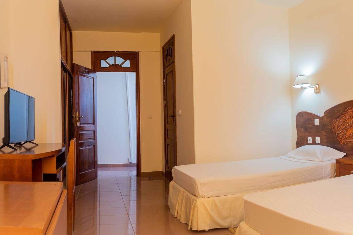 A hotel room with two single beds, a wooden desk, a television, and a door leading to another room.