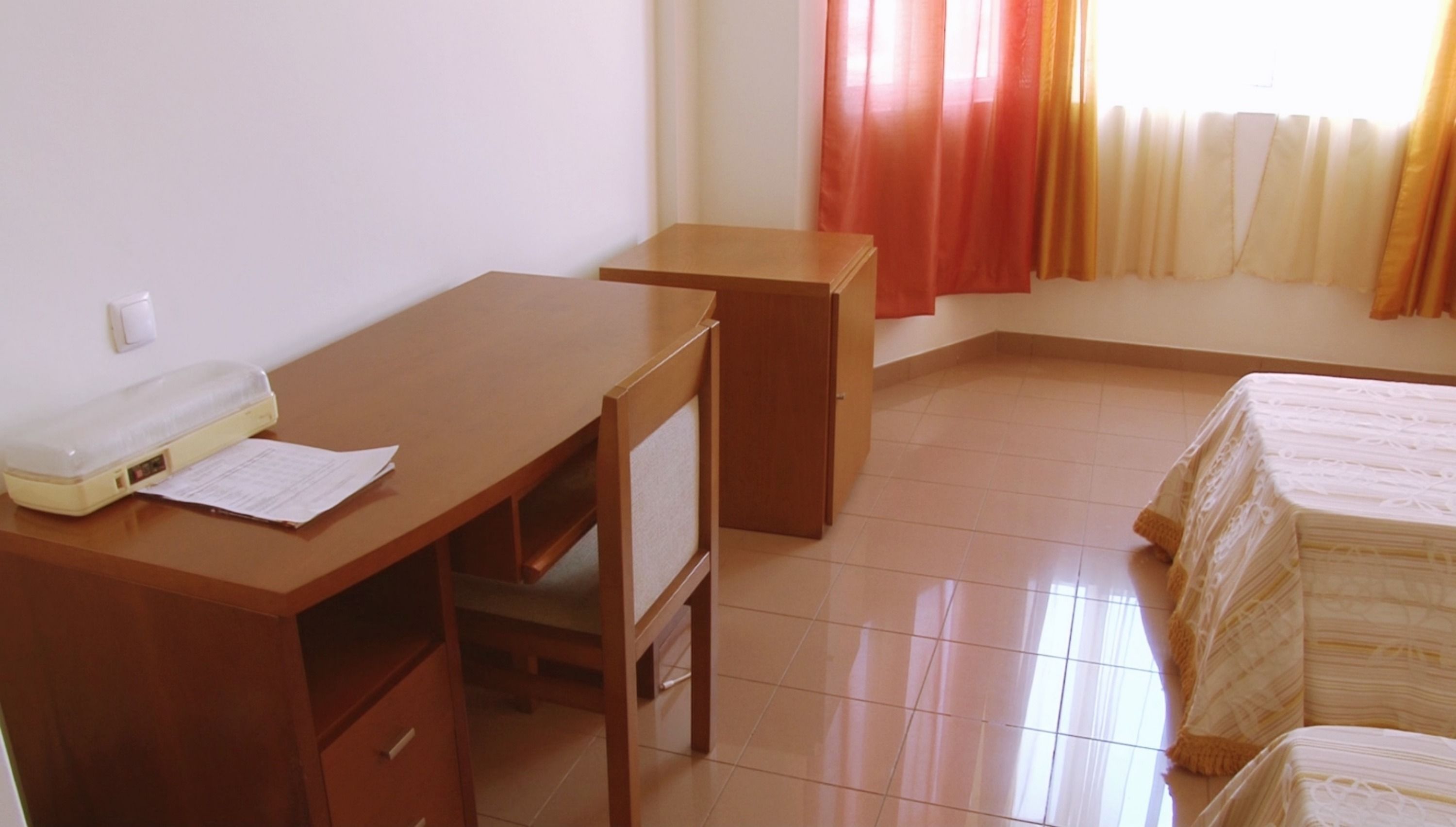 Une pièce comprenant un bureau en bois, une chaise, un lit recouvert d’une courtepointe blanche, un radiateur et une fenêtre avec des rideaux.