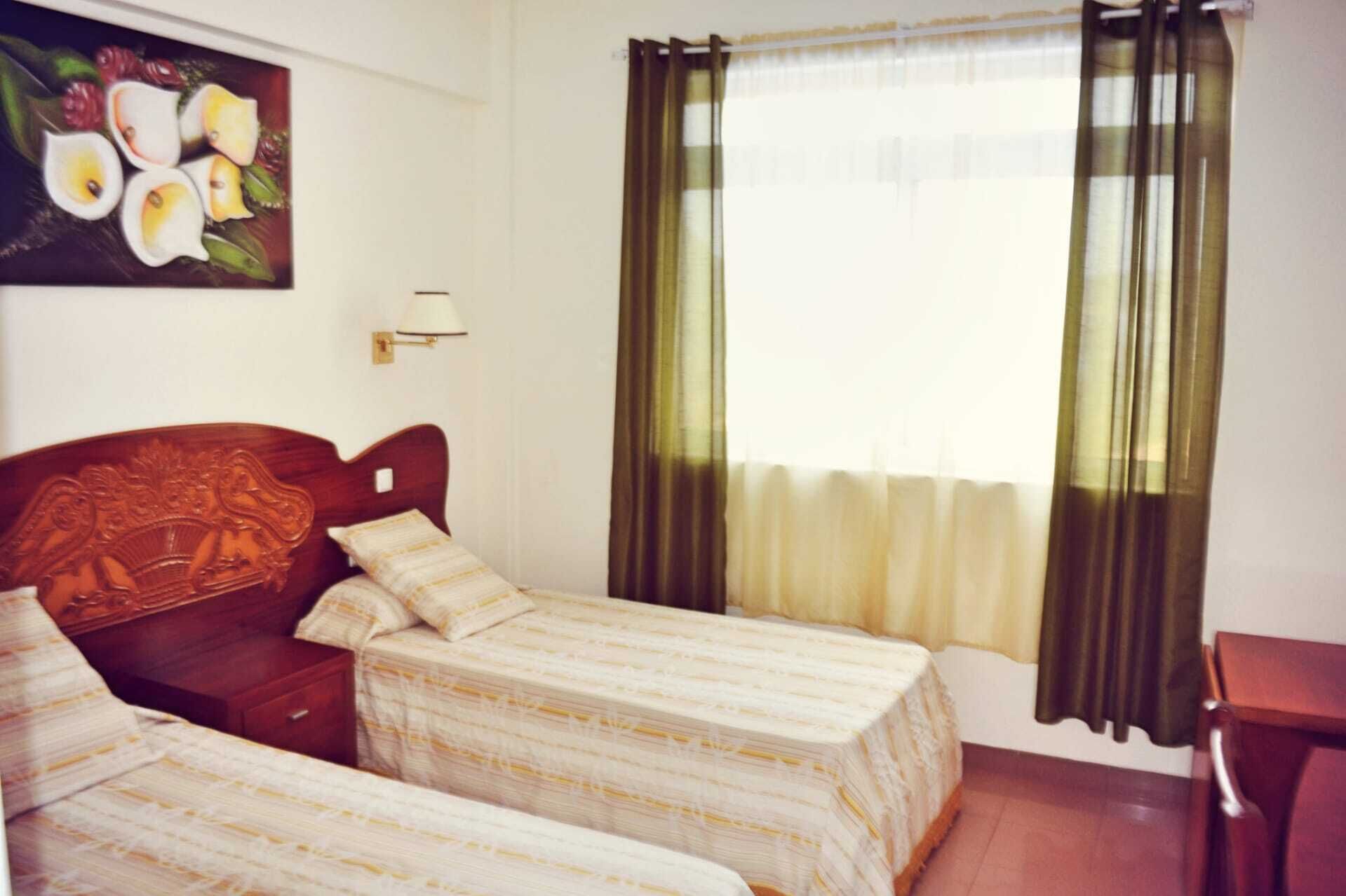 Una habitación con dos camas, un cabecero de madera, una lámpara de pared y un ventanal con cortinas.