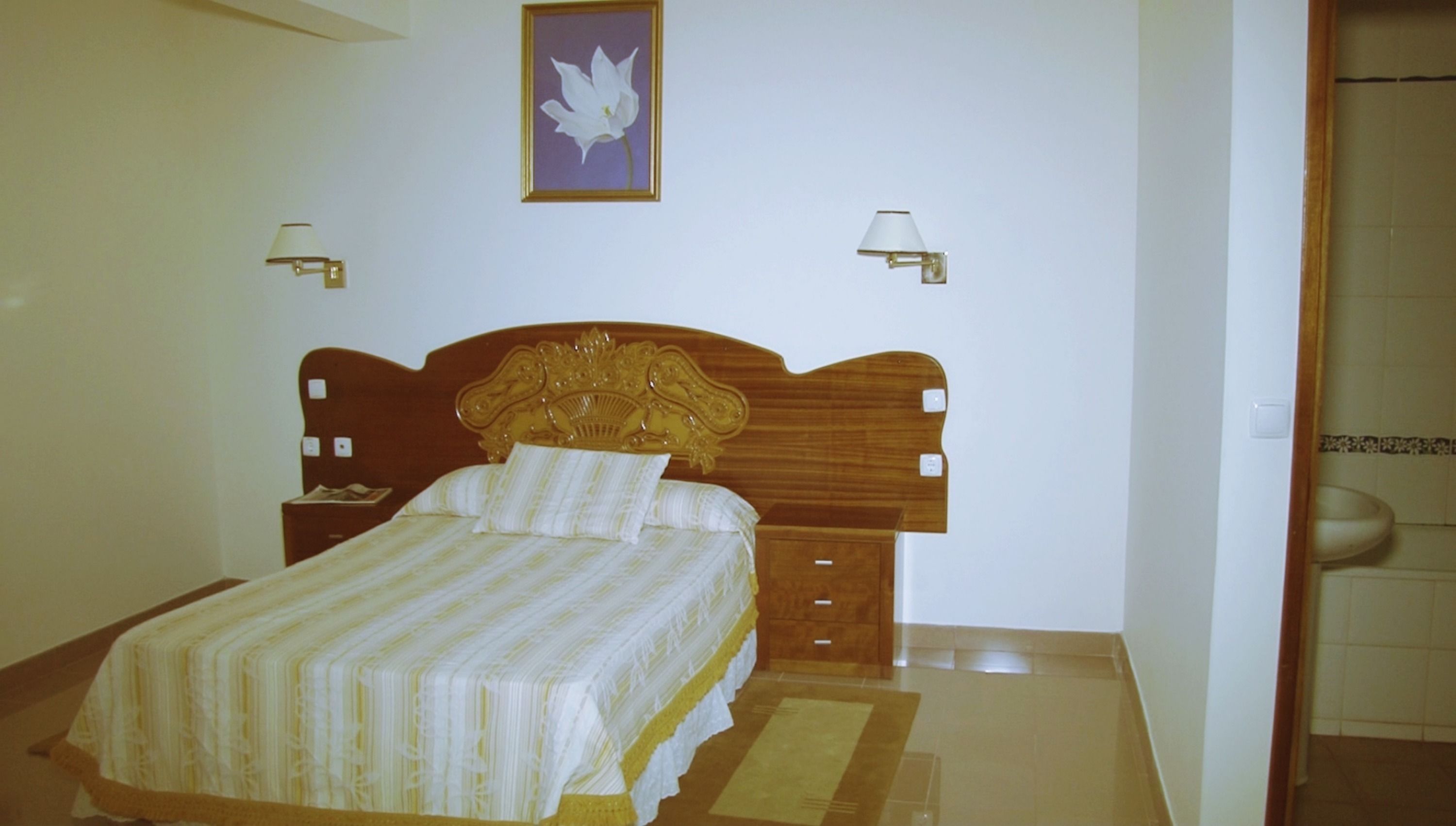 Un dormitorio con cabecera de madera, cama con colcha a rayas, dos mesitas de noche, una lámpara de pared y un cuadro enmarcado en la pared.