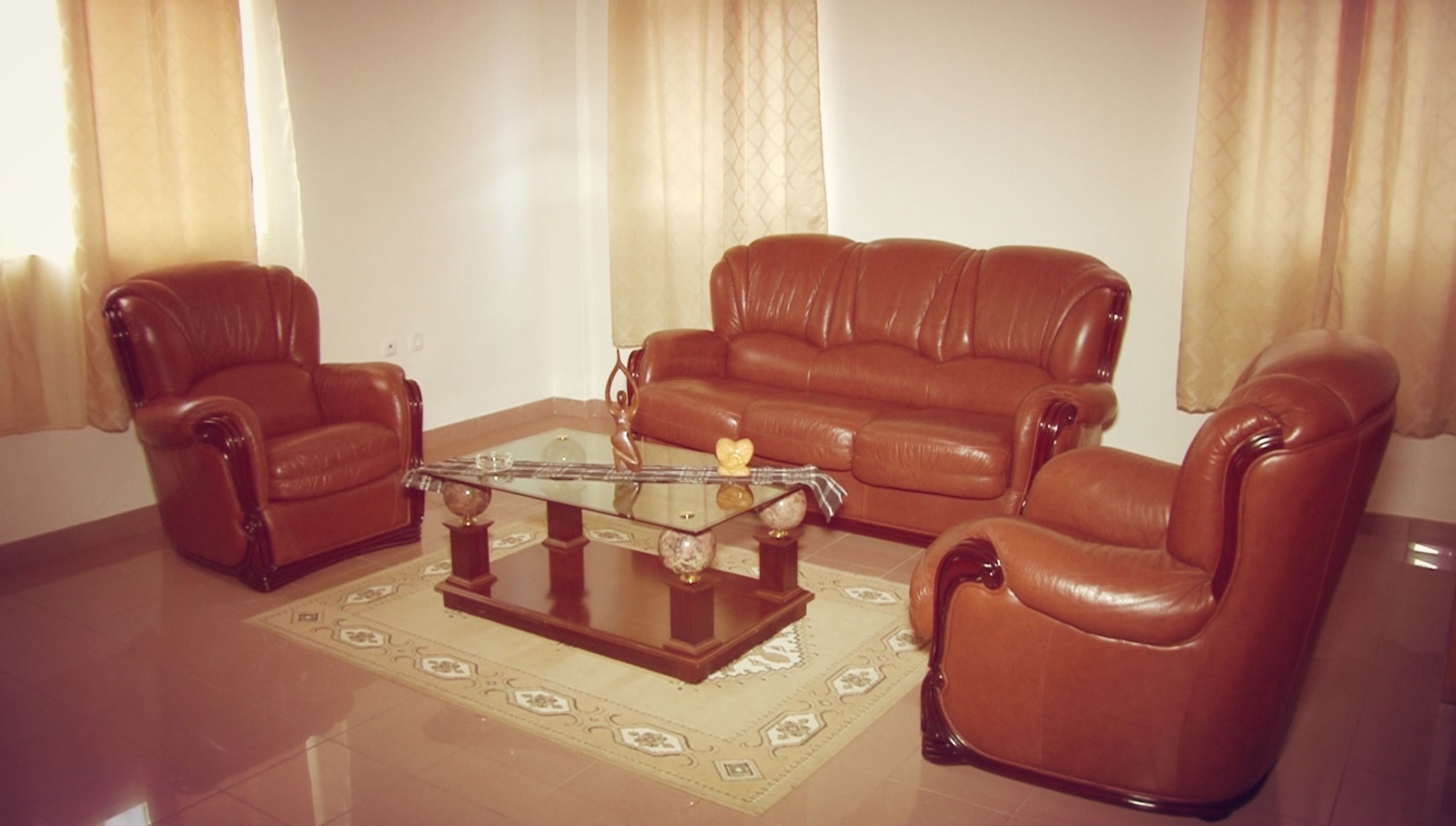 Una sala de estar con un sofá de cuero marrón, dos sillones y una mesa de centro de vidrio con objetos decorativos.
