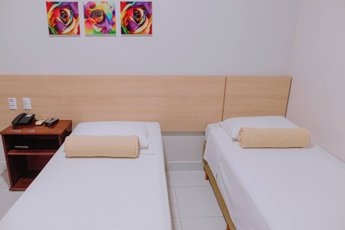 Habitación de hotel con dos camas, una mesita de noche de madera con teléfono y control remoto, y tres cuadros coloridos en la pared.