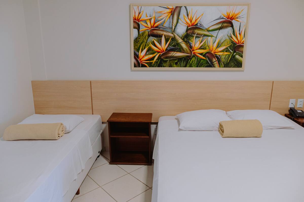 Habitación de hotel con dos camas, cabecero de madera, mesita de noche y un cuadro de flores tropicales en la pared.