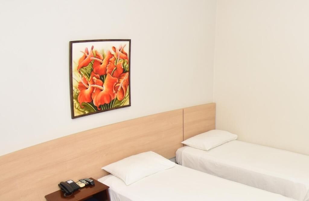 A hotel room with two single beds, a wooden headboard, a bedside table with a phone and a remote control, and a framed picture of flowers on the wall.