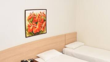 A hotel room with two single beds, a wooden headboard, a bedside table with a phone and a remote control, and a framed picture of flowers on the wall.