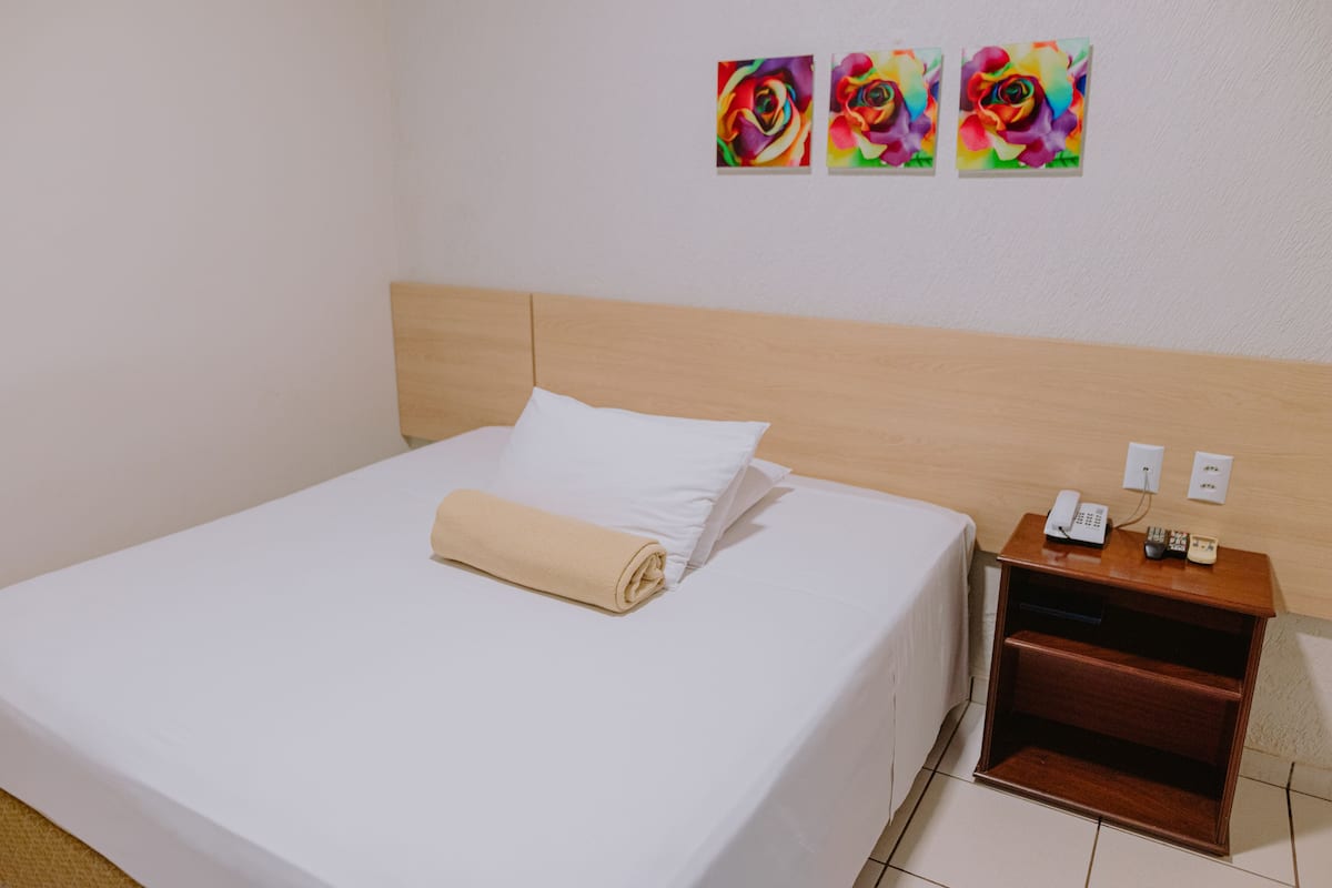 Una cama bien hecha con sábana blanca y toalla color beige, cabecera de madera, mesita de noche con teléfono y control remoto, y tres cuadros de flores coloridas en la pared.