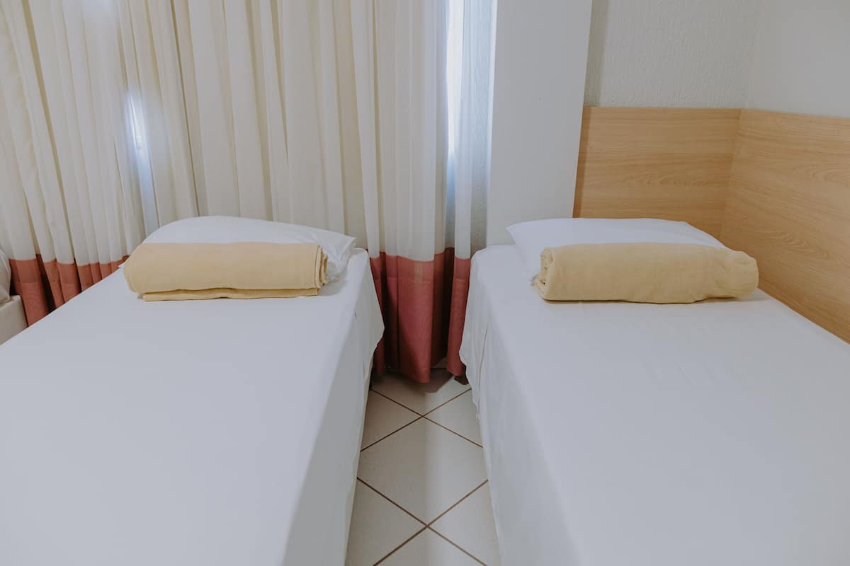 Dos camas individuales con ropa blanca y toallas color beige, un cabecero de madera y piso de baldosas.