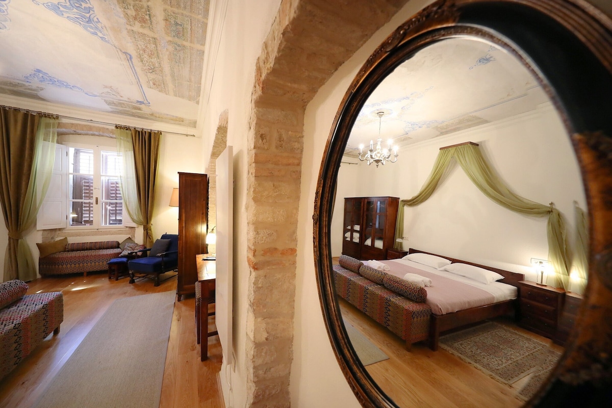 A bedroom with a large mirror reflecting the room, a bed with a patterned bedspread, a wooden wardrobe, and a chandelier.