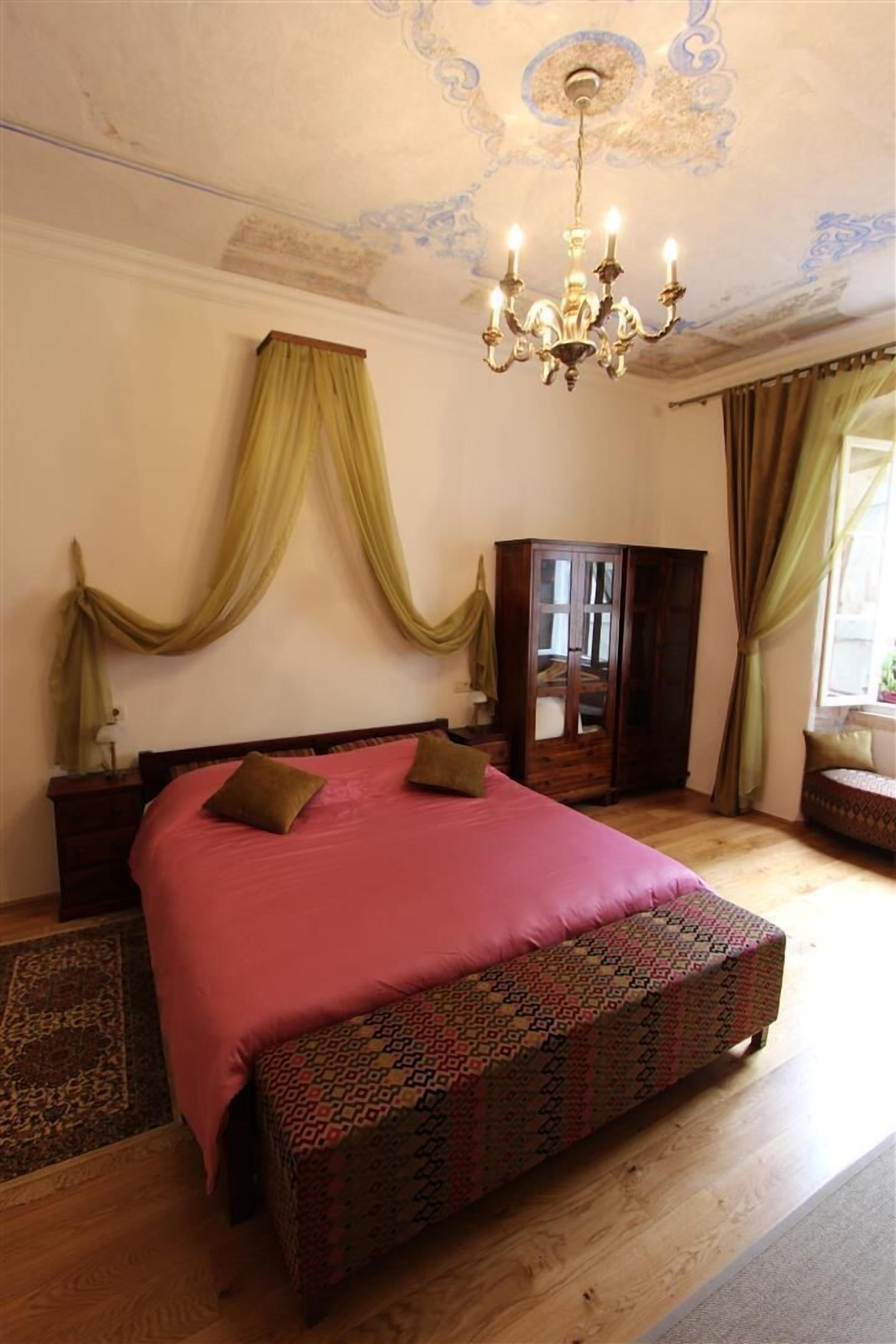 A bedroom with a large bed, a wooden wardrobe, a chandelier, and draped curtains.