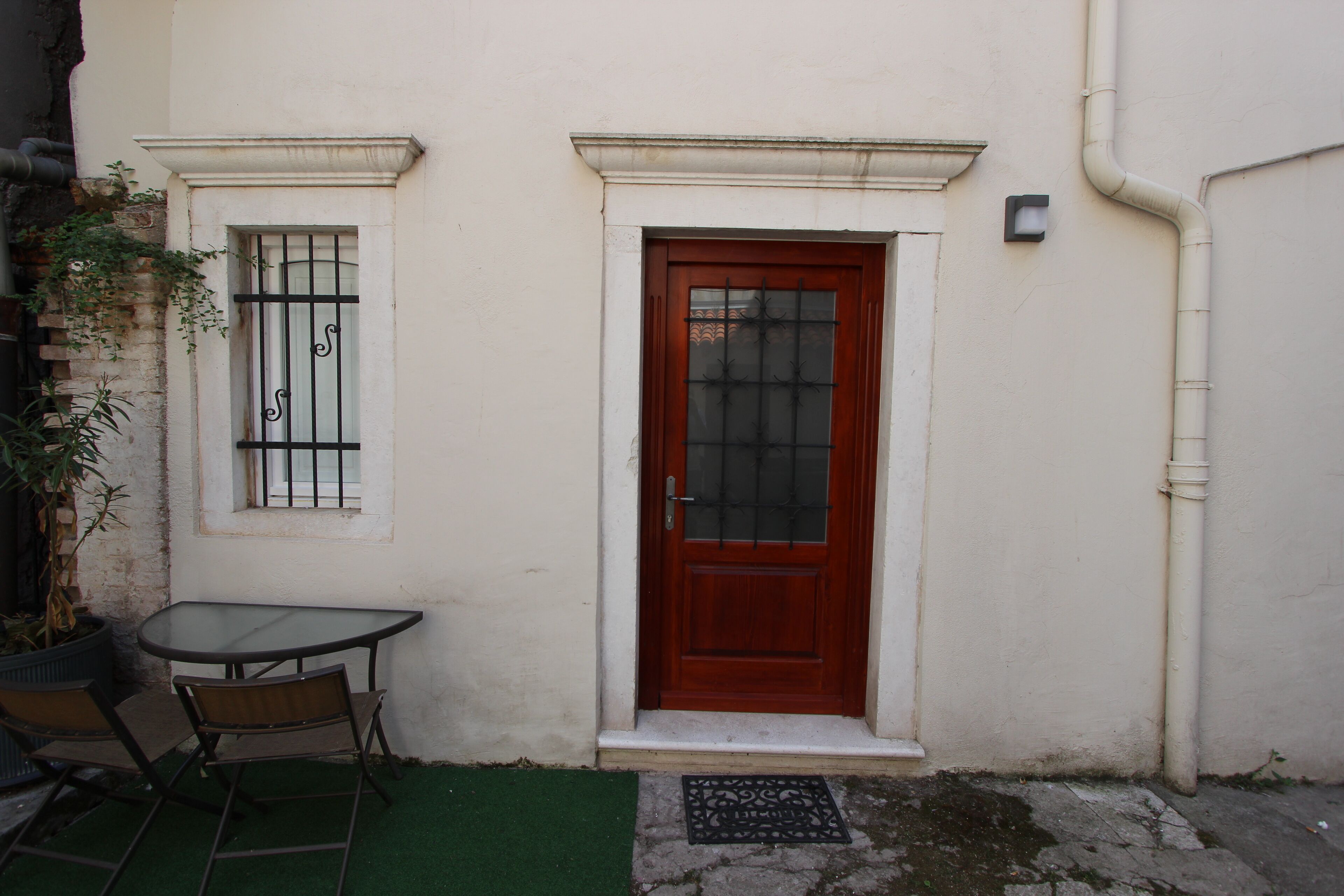 A building with a red door, a window with metal bars, a small table with chairs, and a potted plant.