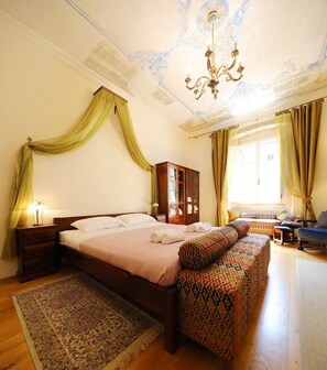 A bedroom with a large bed, a wooden wardrobe, a bench with patterned cushions, a chandelier, and a large window with curtains.