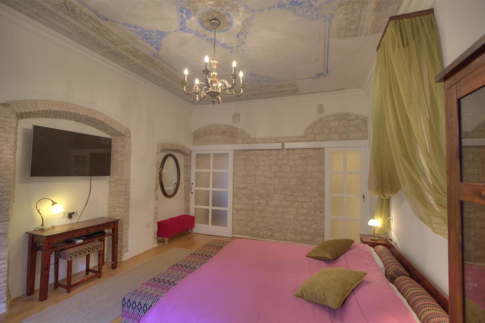 Une chambre à coucher avec un grand lit, un lustre, une télévision, un banc et une porte à panneaux de verre.