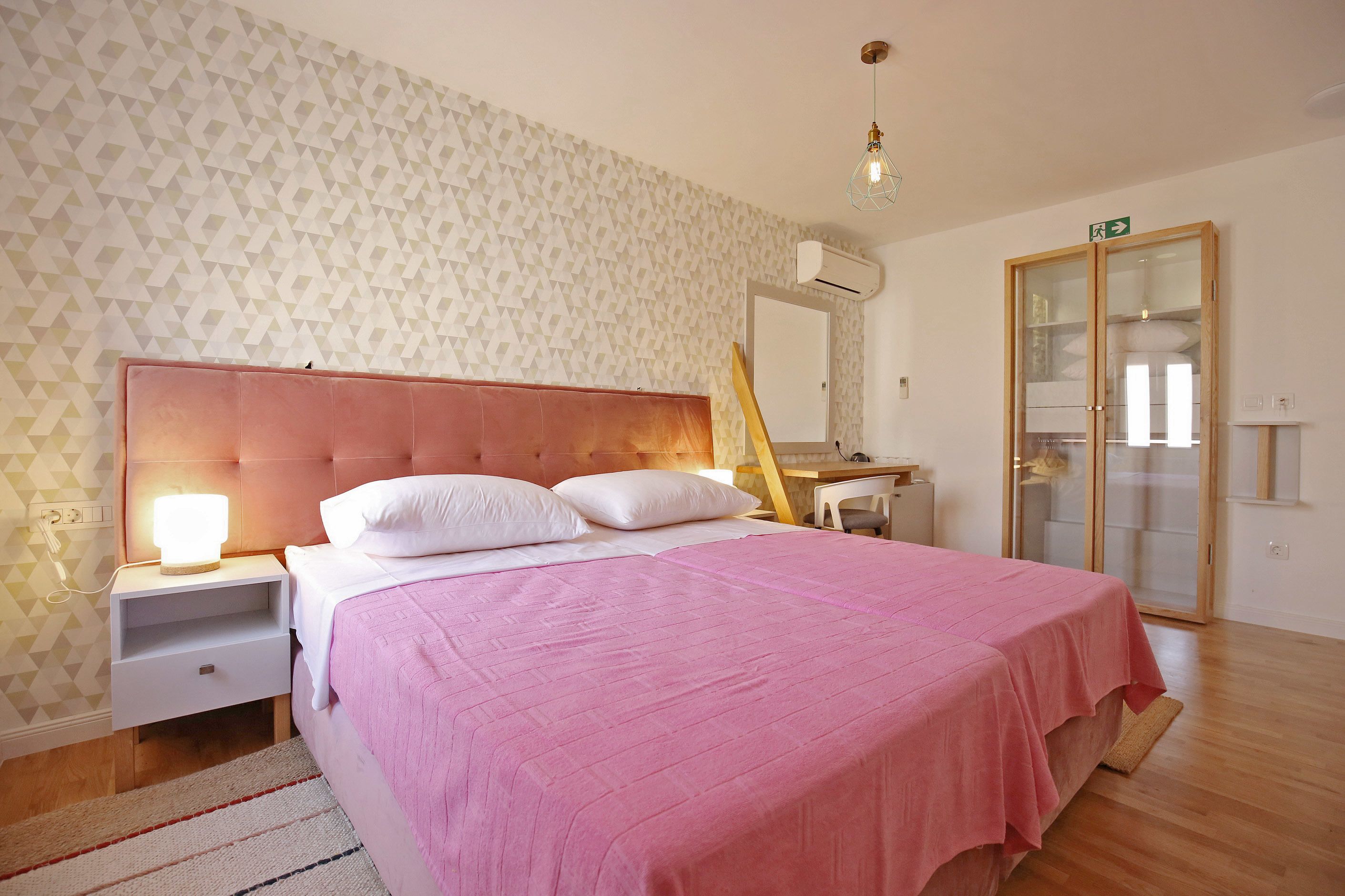Un dormitorio con colcha rosa, cabecera de madera, lámpara de noche, mesita de noche, espejo y armario.