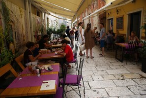 An outdoor cafe with wooden tables and purple chairs, surrounded by stone walls and potted plants.