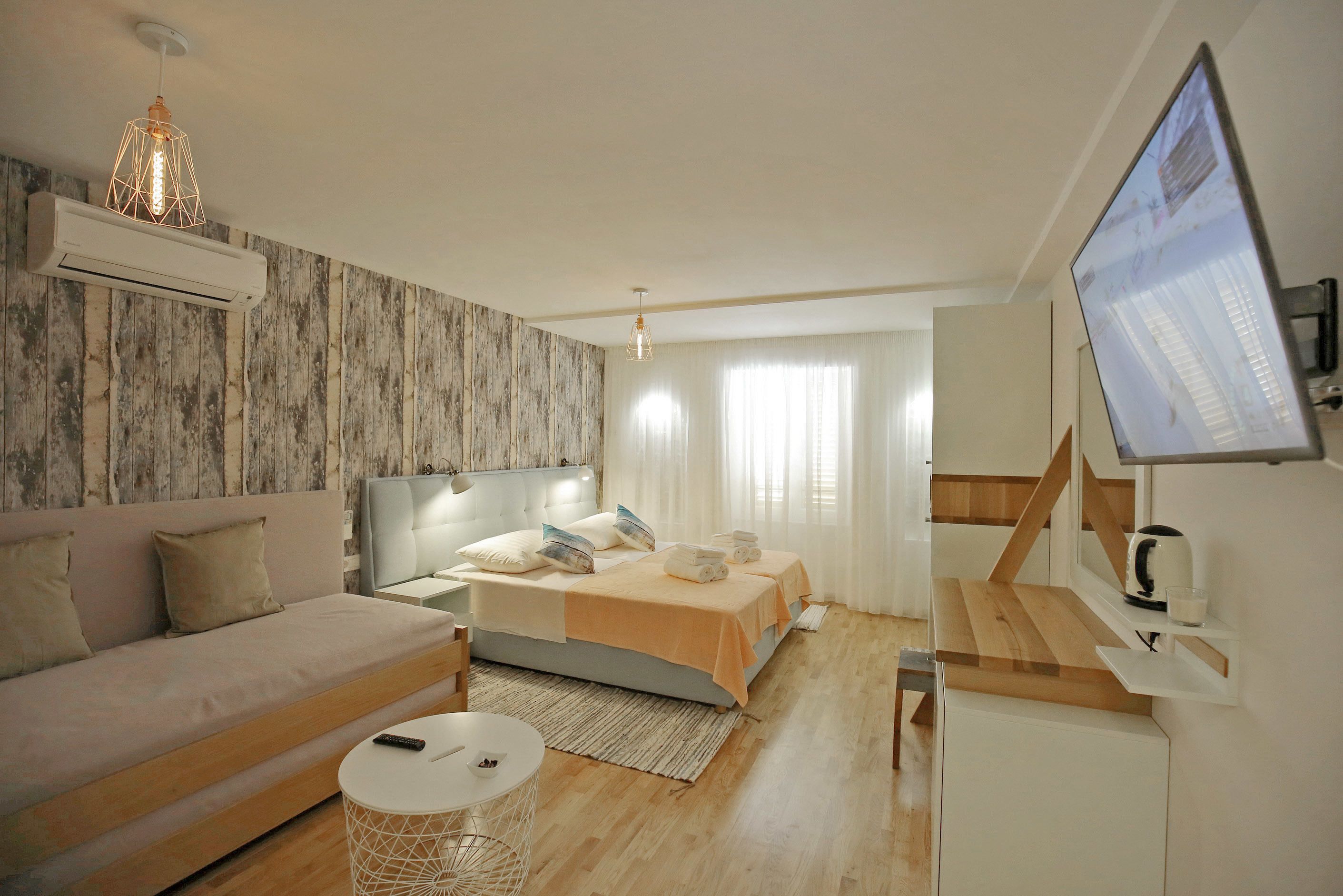 A modern hotel room with a sofa, a bed with two pillows, a bedside table, a TV, and a wooden floor.