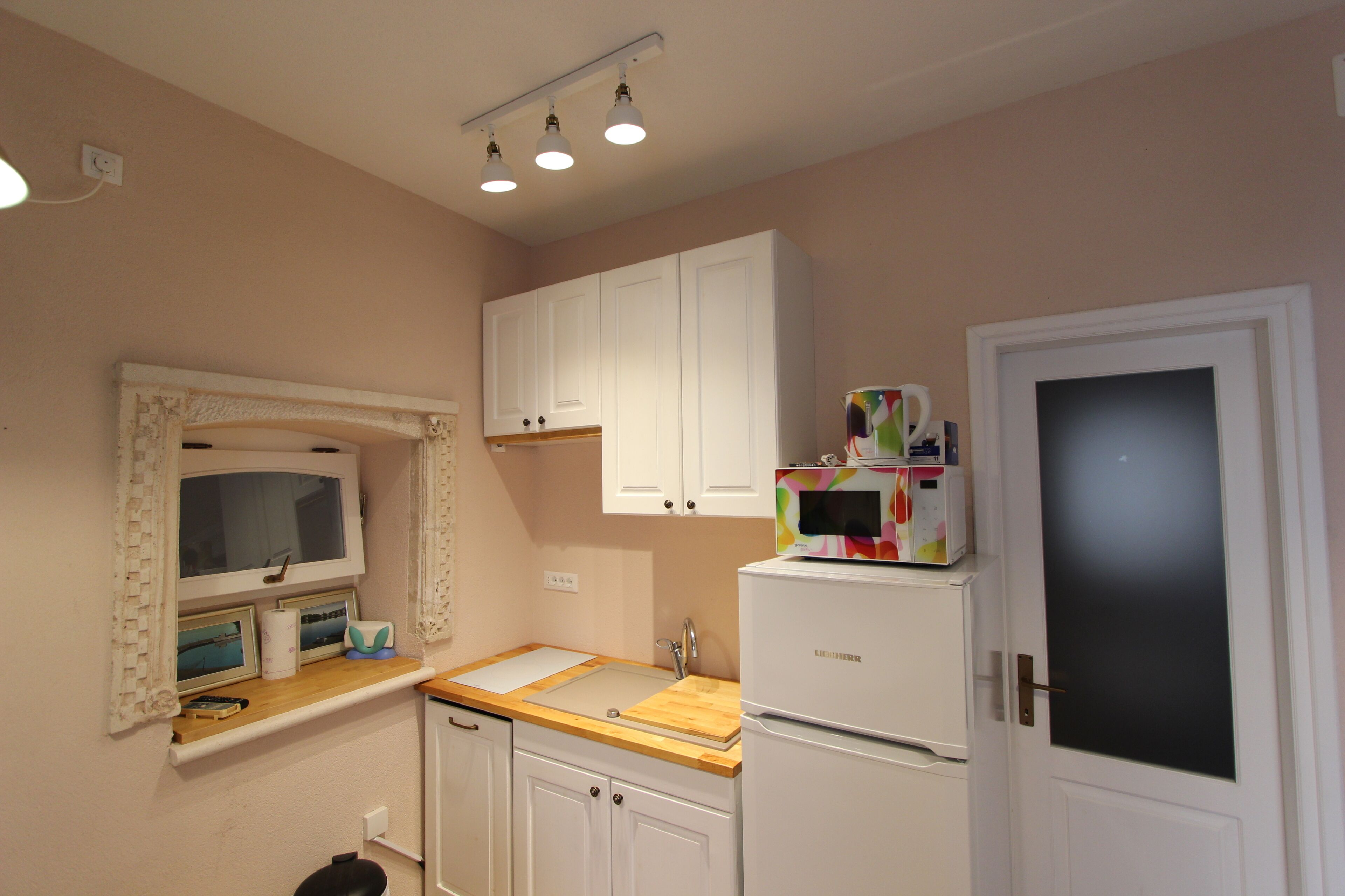 A small kitchen with white cabinets, a microwave, and a refrigerator. There is a window with a curtain and a shelf with pictures and a tissue box.