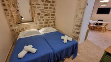 Una habitación con pared de piedra, cama con ropa de cama azul, puerta blanca y una pequeña mesa con sillas.
