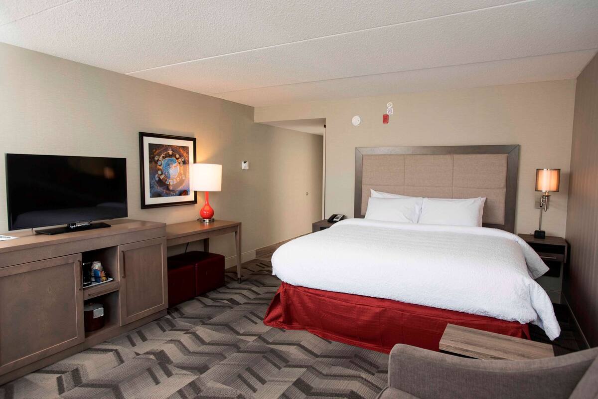 Ein Hotelzimmer mit einem großen Bett, einem Fernseher, einem Schreibtisch mit Lampe und einem roten Teppich.