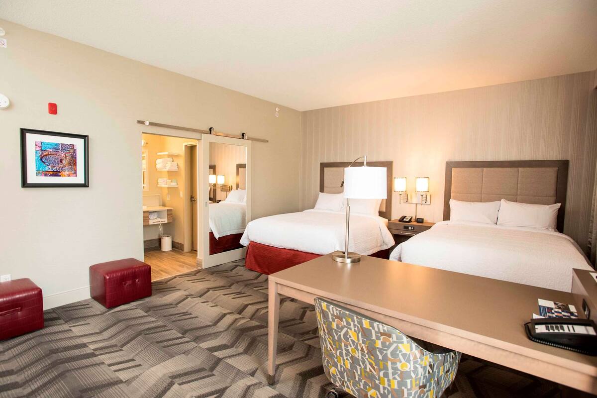 Ein Hotelzimmer mit zwei Betten, einem Schreibtisch, einem Sessel, einem roten Hocker und einem durchgegangenen Türspalte sichtbaren Badezimmer.