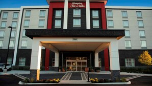 Fachada de un hotel Hampton Inn & Suites con diseño moderno, que destaca por su entrada principal, el número '760' sobre las puertas y un cielo despejado al fondo.