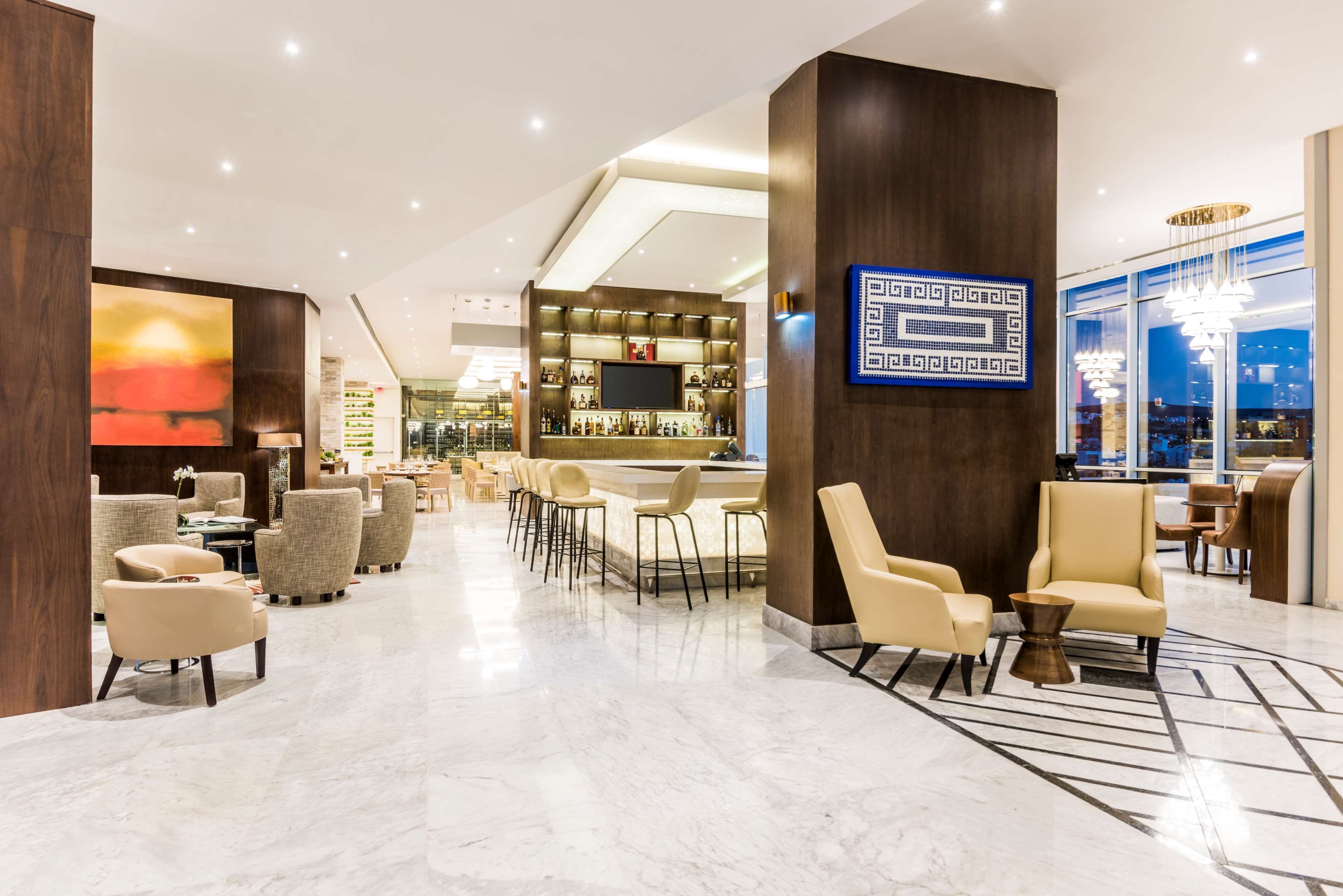 Un hall d’hôtel moderne doté d’un bar, d’espaces aménagés avec des sièges et d’un grand écran numérique.