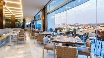 Un comedor moderno con mesas y sillas, ventanas amplias que ofrecen vista a la ciudad, y un rincón de estar con sofá.