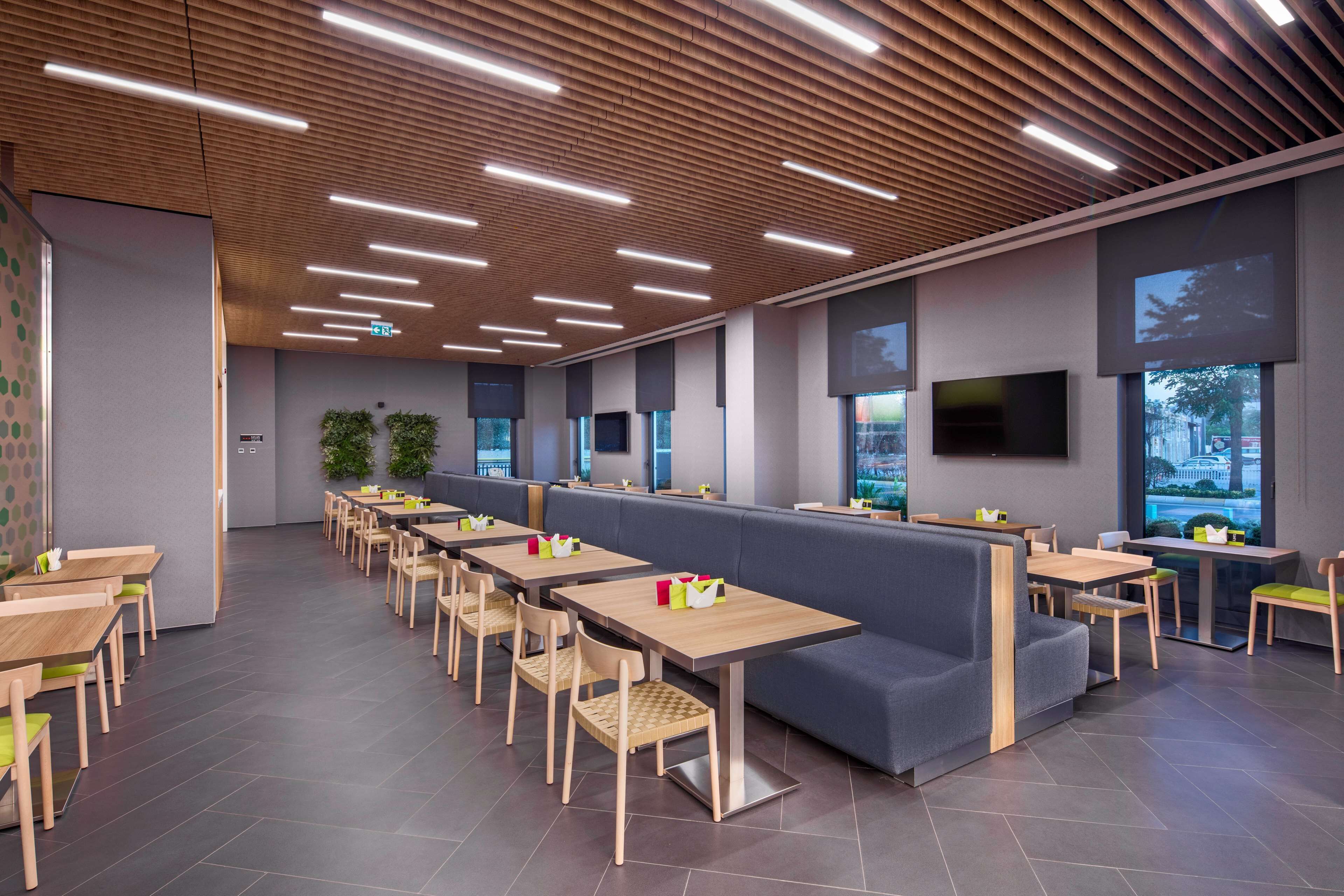 A modern restaurant with wooden tables and benches, large windows, and a flat-screen TV mounted on the wall.
