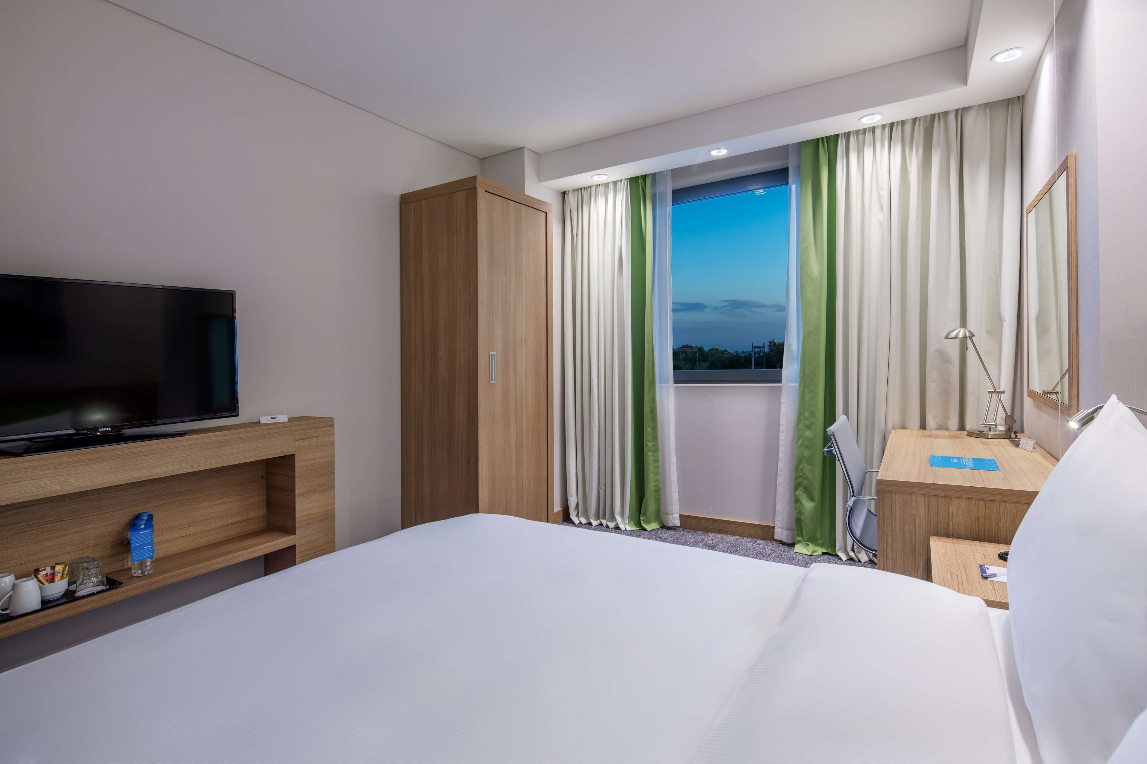 Ein Hotelzimmer mit einem großen Bett, einem Fernseher auf einer Holzablage, einem Schreibtisch mit einer Lampe und Blick auf die Stadt durch das Fenster.