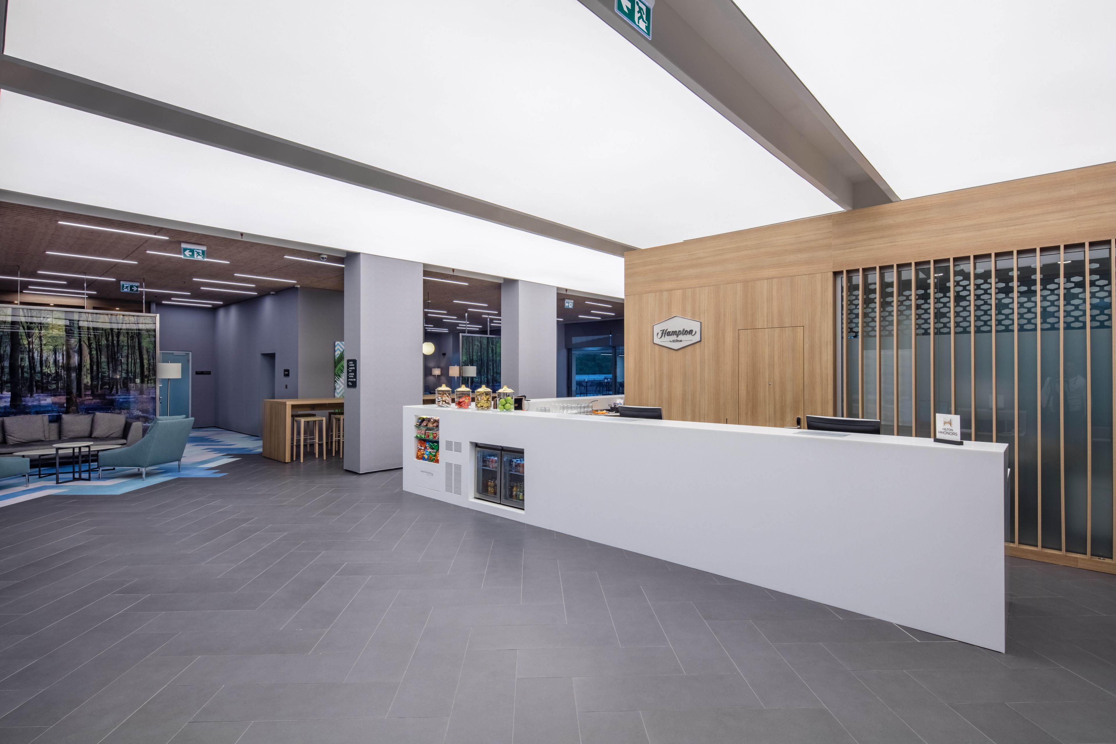Un espace d’accueil moderne, avec un comptoir de réception blanc, un coin salon comprenant un canapé et des fauteuils, et un mur décoratif en bois agrémenté d’une enseigne.