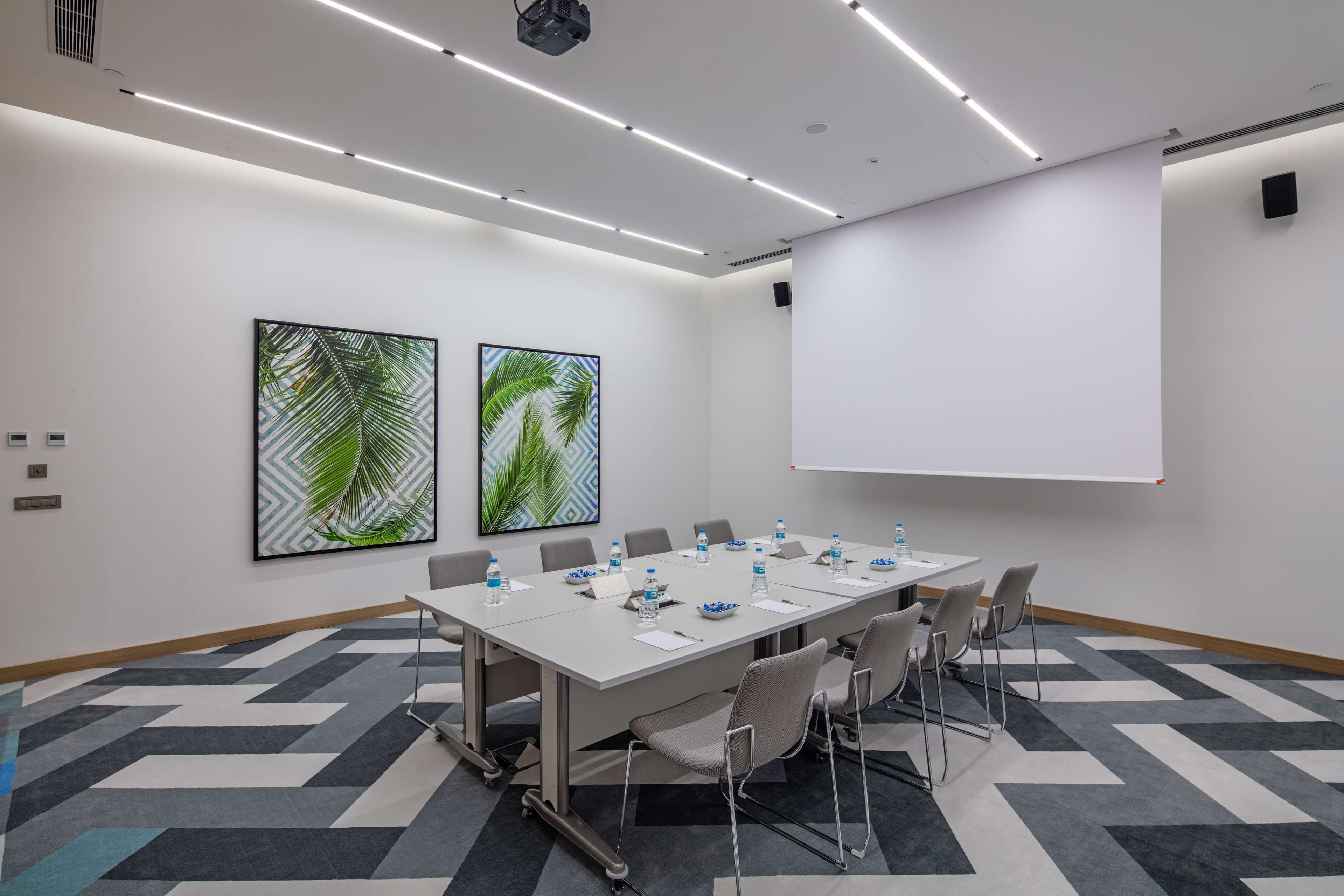 Ein Konferenzraum mit langem Tisch, Stühlen, Wasserflaschen und einer Projektionsleinwand.