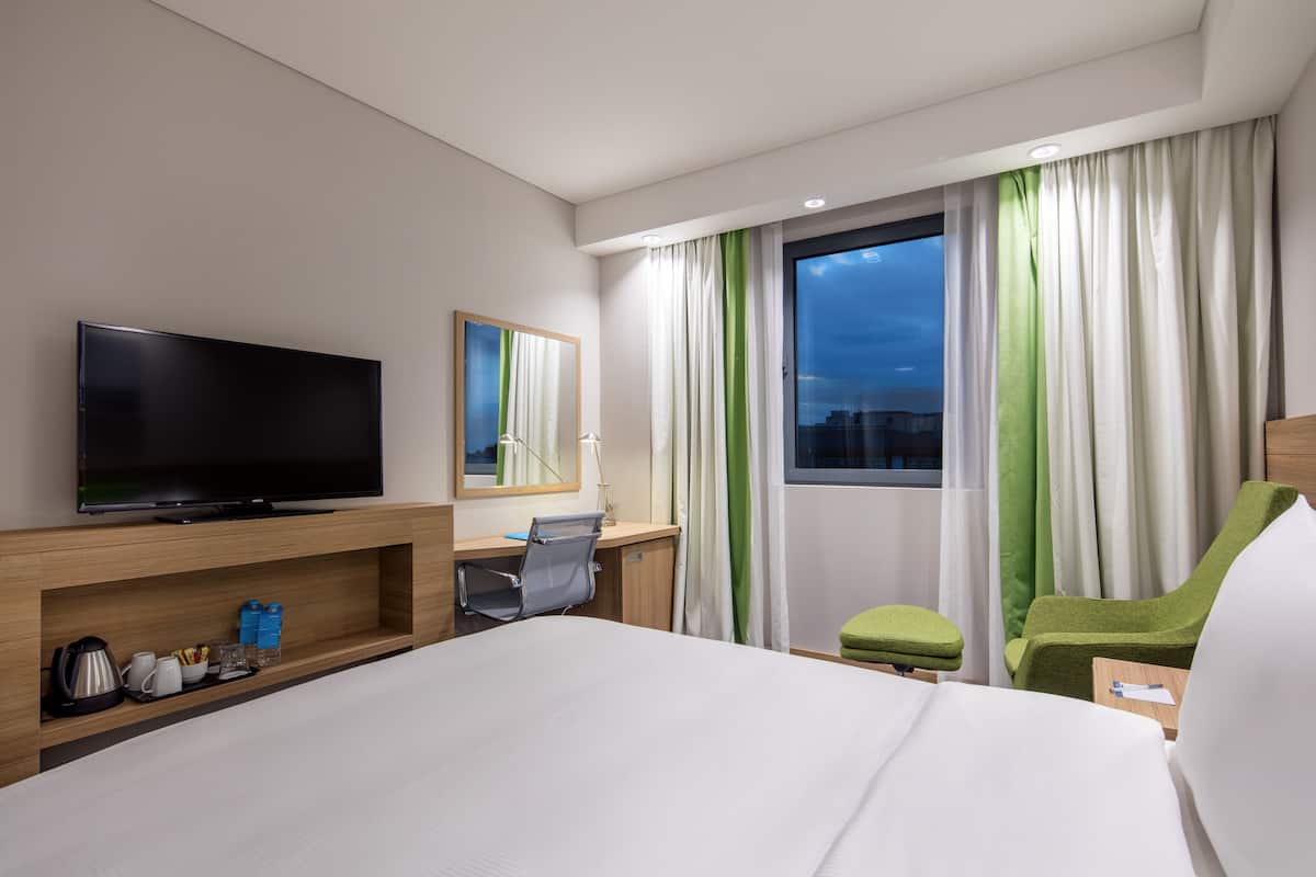 Une chambre d’hôtel avec un lit, un bureau équipé d’un ordinateur, une télévision et une fenêtre avec des rideaux.