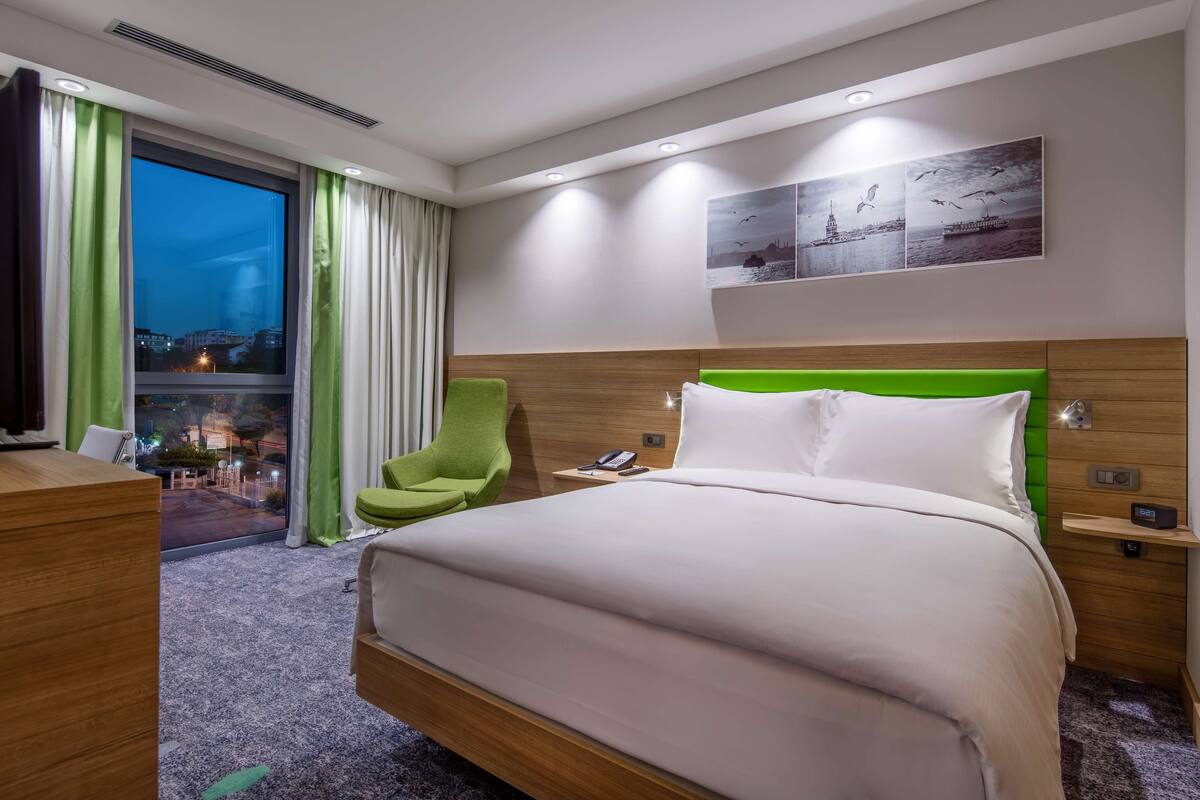 Une chambre d’hôtel avec un grand lit, un fauteuil vert, un bureau et une vue sur la ville la nuit.