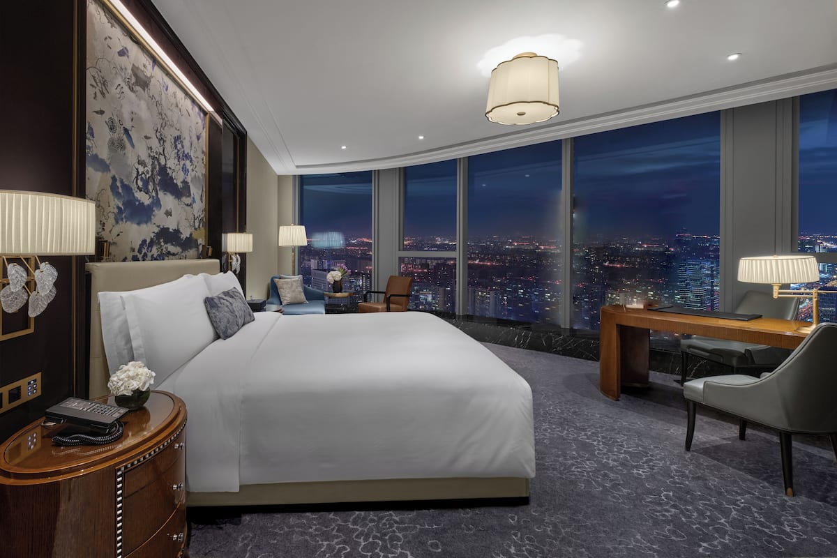 A hotel room with a large bed, a desk, a chair, and a view of the city at night.
