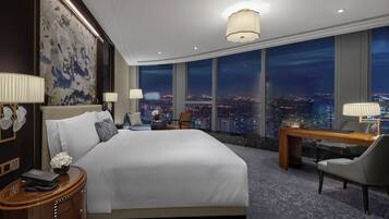 A hotel room with a large bed, a desk, a chair, and a view of the city at night.