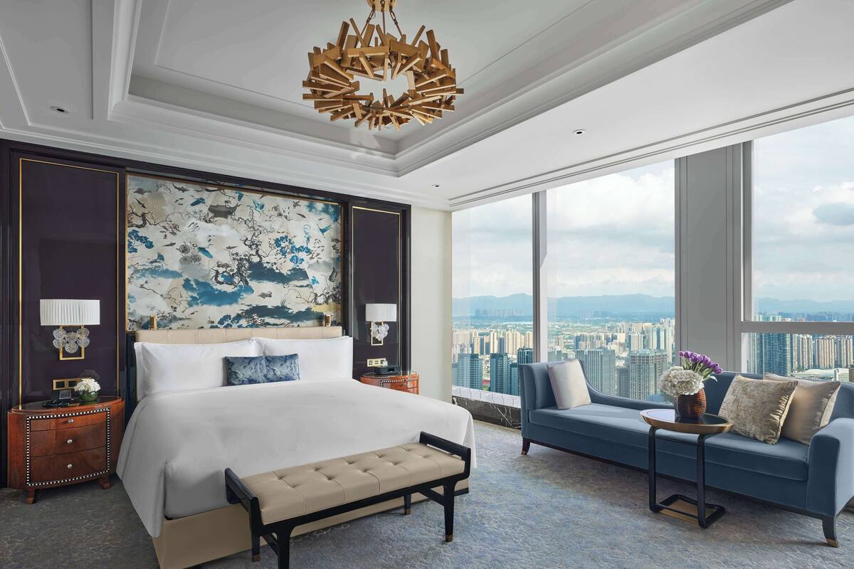 A modern hotel bedroom with a large bed, a blue sofa, and a city view.