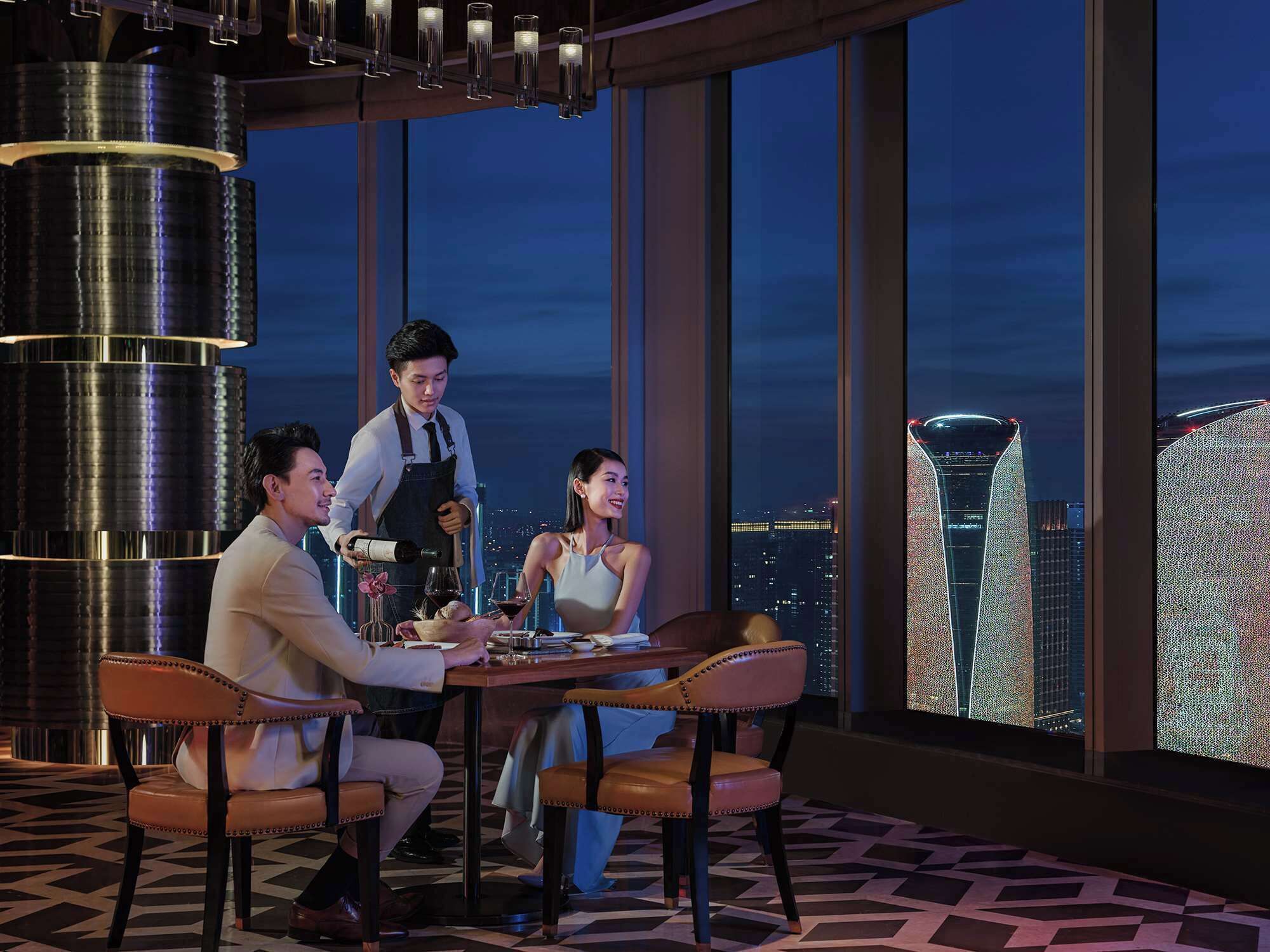 A high-rise restaurant with a panoramic city view, a waiter serving, and a table set for three.